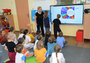 Dzieci siedzą w grupce przed monitorem interaktywnym, dwoje dzieci uruchamia program "Dobre uczynki"